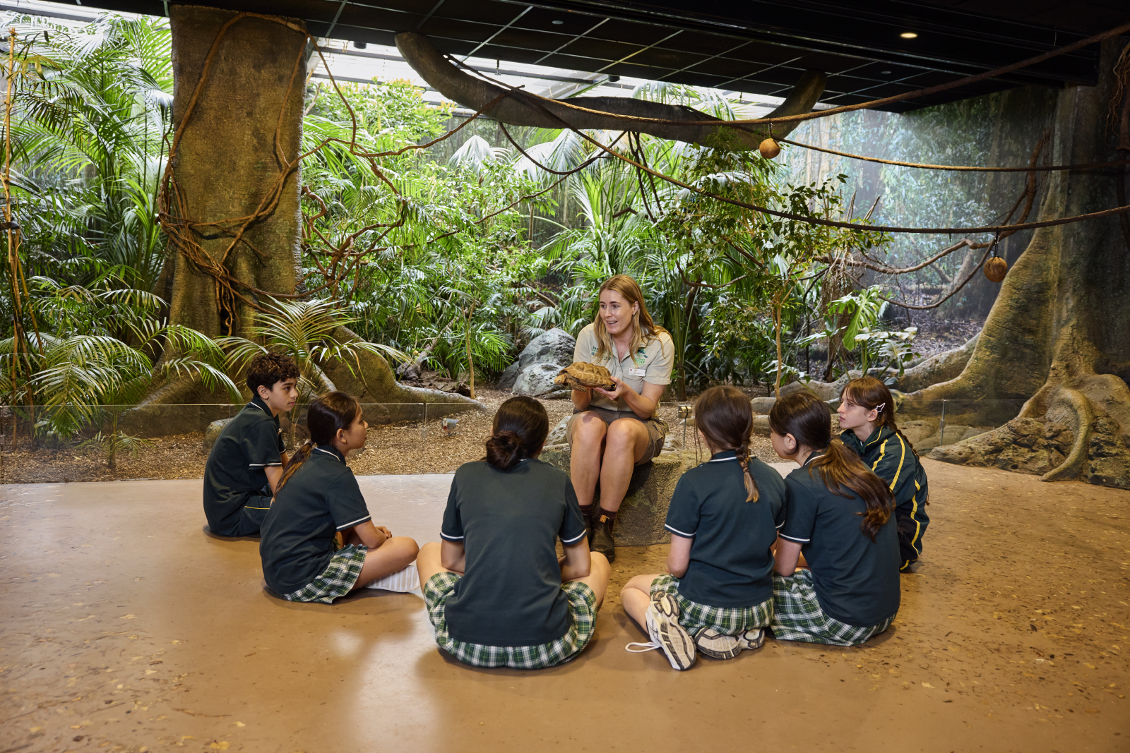 Children in a classroom listening to a teacher with greenery in background. 