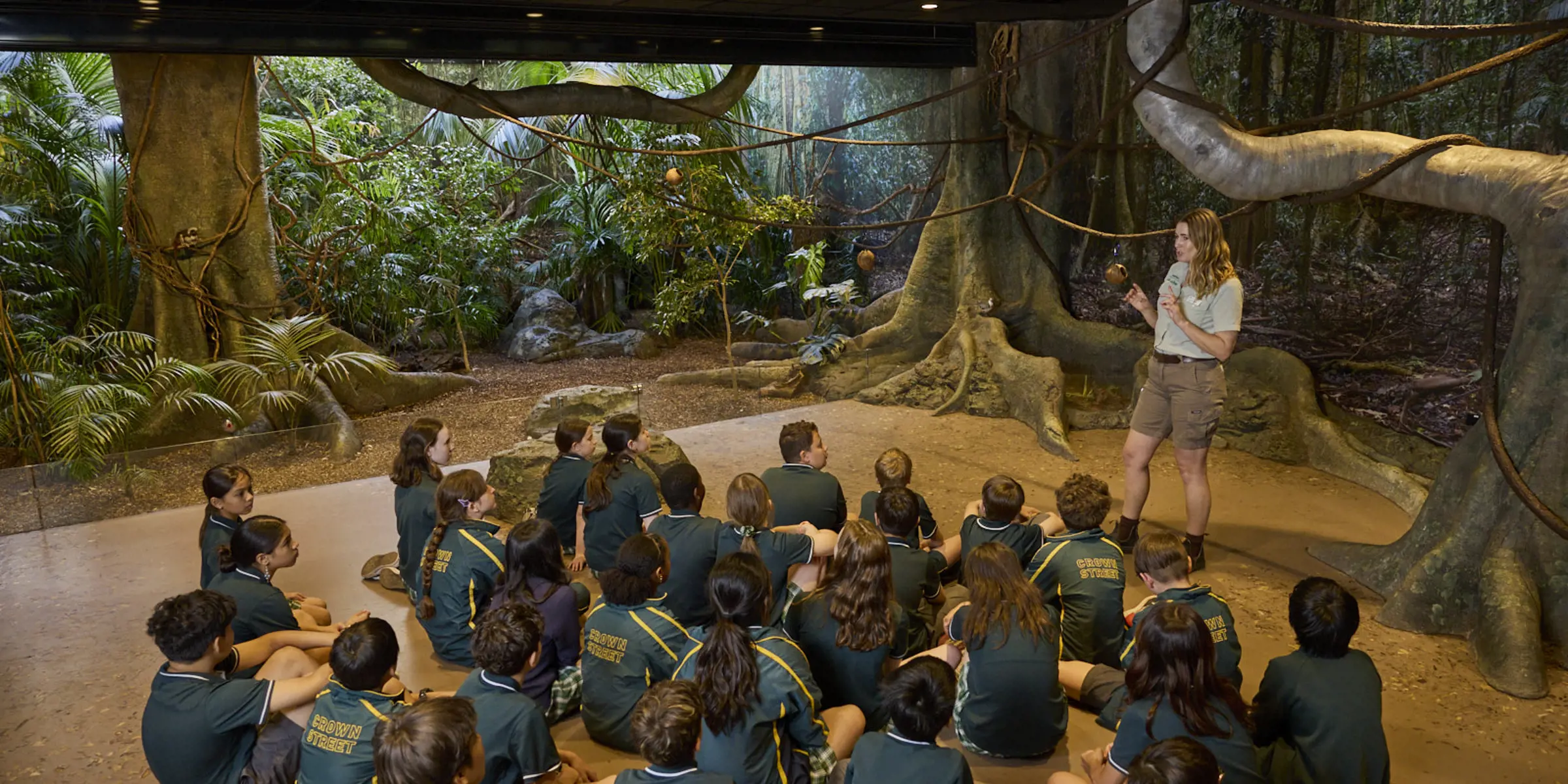 Students seated indoors listening to a teacher in a rainforest-themed immersive classroom.