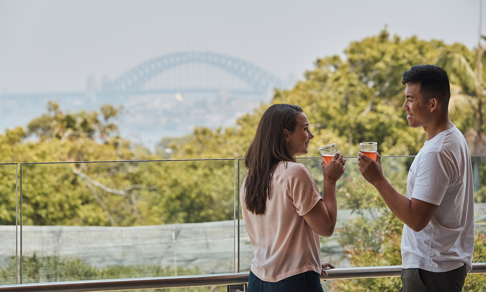 Couple enjoying a drink on the balcony of the N'Gurra Lounge overlooking Sydney Harbour at the Wildlife Retreat at Taronga.